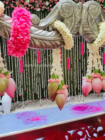 A close-up of a silver cradle decorated with hanging lotus buds and pink floral garlands. The backdrop of hanging jasmine strings adds a delicate and fragrant touch to the setup.
