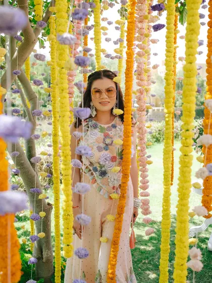 The bride poses amidst a curtain of hanging flower garlands in shades of yellow, orange, and purple, creating a perfect photo moment.