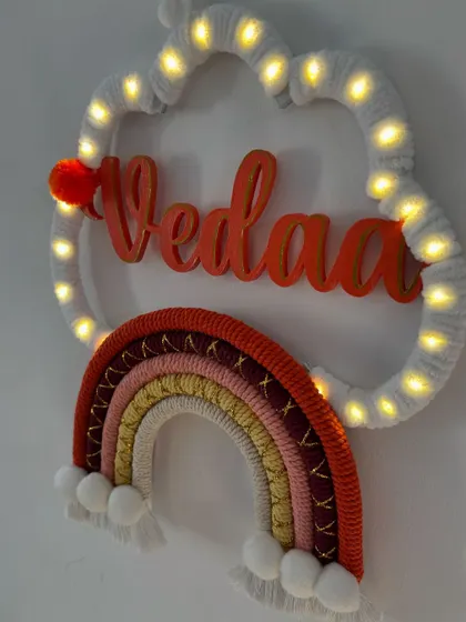 The 'Vedaa' name hanging glowing beautifully with fairy lights. This piece uses a warm, sunset-inspired color palette for the rainbow.