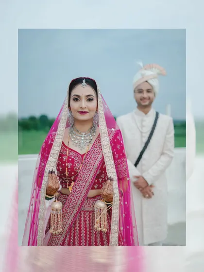 A beautiful portrait of a North Indian bride in her pink lehenga, with the groom standing softly out of focus in the background.