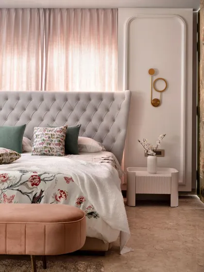 This master bedroom is designed as a soft, classical retreat with a tufted wingback bed and a custom floral bedspread. The color palette is a gentle mix of pastels, with panel moulding and a unique brass wall sconce adding a touch of elegance.