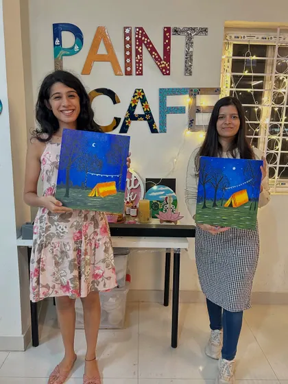 Two participants with their finished canvas paintings of a campsite at night.