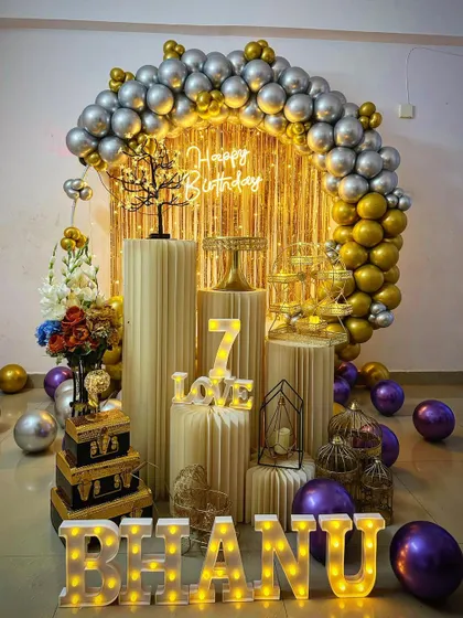 A 7th birthday celebration with a gold and silver theme. The setup includes a balloon arch, shimmer curtain, neon sign, and props, with purple balloons scattered on the floor for an extra pop of color.