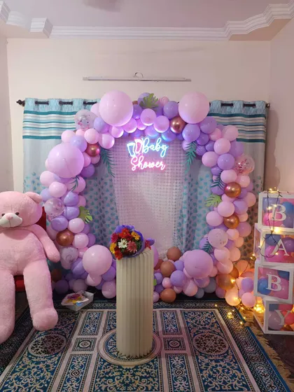 A cozy baby shower setup in a living room. A circular balloon arch in pastel purple and pink frames a mesh backdrop, with a large teddy bear and 'BABY' blocks.