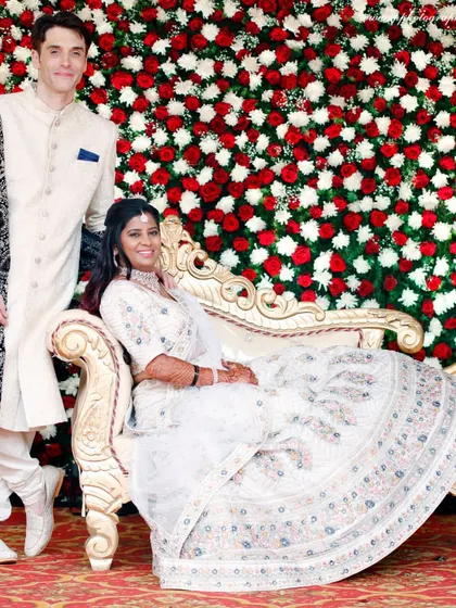 A stunning portrait of the couple against a wall of red and white roses. The vibrant backdrop makes their elegant white outfits stand out beautifully.