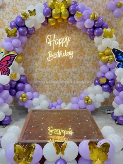 A butterfly-themed balloon ring in purple, white, and gold. The decoration features large butterfly cutouts on the balloon columns and smaller butterflies scattered throughout the ring for a whimsical feel.
