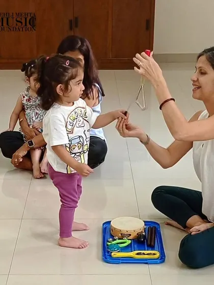 A teacher introduces a toddler to the triangle, a simple percussion instrument. Early exposure to different instruments helps develop a child's listening skills and curiosity.