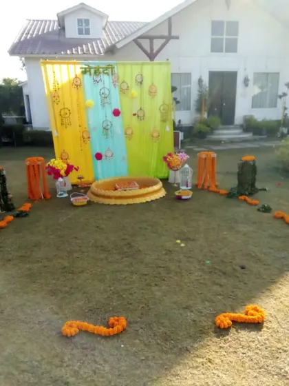 A simple outdoor Mehendi setup in a lawn, featuring a colorful backdrop and a central 'urli' for rituals.