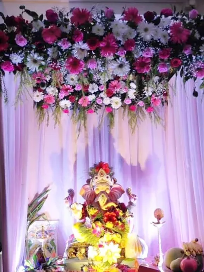 A beautiful hanging floral arrangement of pink and white gerberas and daisies creates a canopy effect over the Ganpati idol. The soft purple lighting enhances the festive mood.