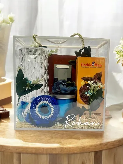 This personalized Rakhi hamper for Rohan is presented in a clear acrylic bag. It features a crystal-cut vase, a blue evil eye charm, cookies, and other curated items in a cool blue theme.