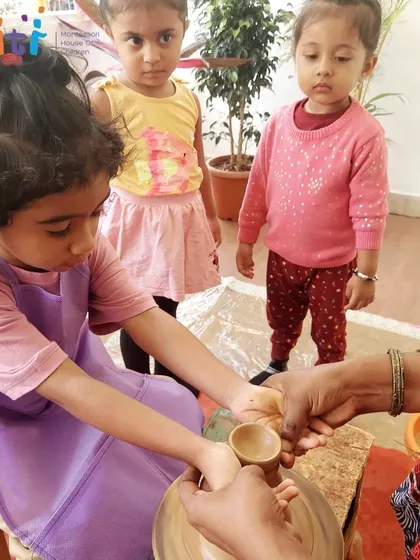 A teacher guides a child's hands on the pottery wheel, helping her form a small clay diya. This one-on-one interaction is a special part of the learning process.
