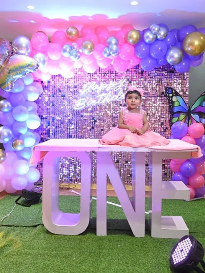 The birthday girl sits proudly on a "ONE" letter table in her butterfly-themed party. This setup includes a pink sequin wall, a balloon arch in shades of pink and purple, and a light-up number '1'.
