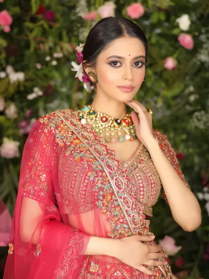 A half-body shot of a model in a pink embroidered lehenga against a floral wall. This image showcases the detailed craftsmanship of the blouse and the colorful statement necklace.