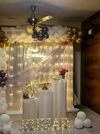An elegant surprise birthday setup perfect for a home celebration. The decor features a white and gold balloon arch, a sheer curtain with fairy lights, and classic white pedestals.
