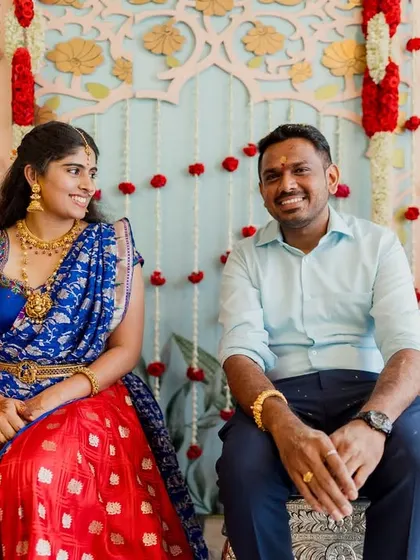 A happy couple seated at their engagement ceremony. The backdrop features a custom-painted panel and hanging floral strings, creating a simple yet beautiful setting.