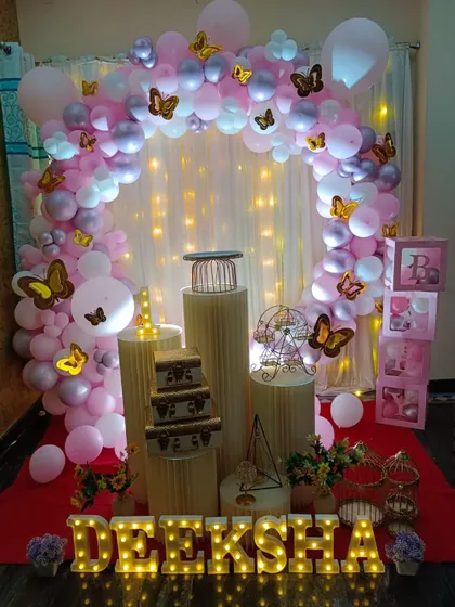 An elegant butterfly-themed birthday setup with a circular balloon arch in shades of pink and silver against a fairy-lit curtain.