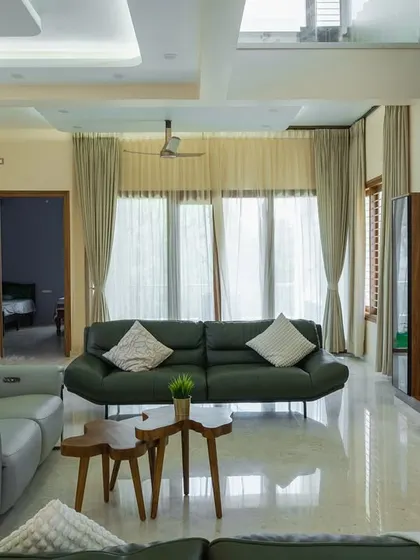 A wide view of a modern villa living room with a double-height ceiling effect. The space features a stylish TV unit, comfortable leather sofas, and large windows that flood the room with natural light.