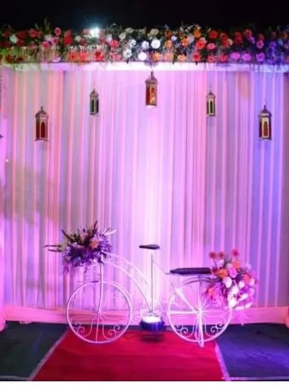 A whimsical photo booth for a reception or sangeet, featuring a white curtain backdrop, a floral arch, hanging lanterns, and a decorative bicycle prop.