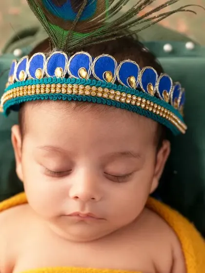 A serene close-up of a sleeping newborn with a Krishna crown and peacock feather.