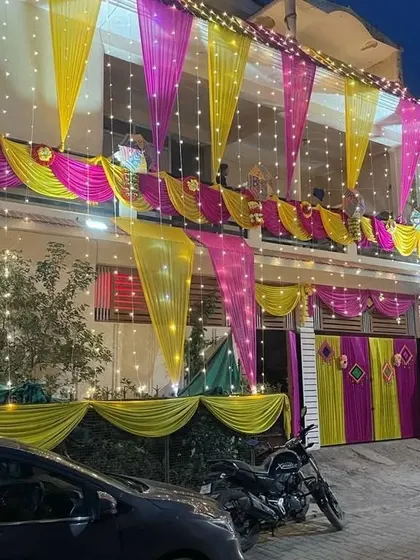 A house decorated for a night function, with yellow and pink drapes on the balcony and gate, all lit up with fairy lights.