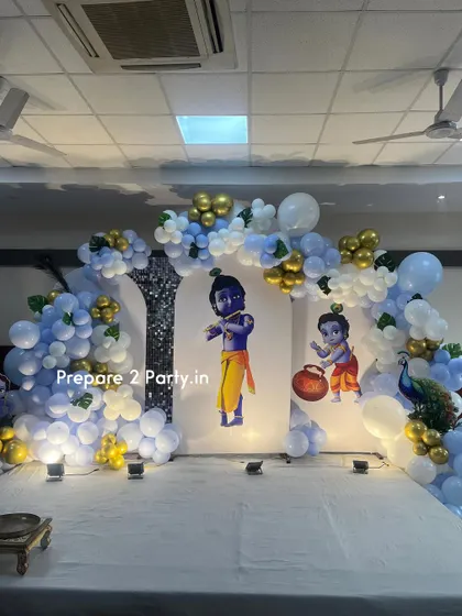 A grand and traditional Krishna-themed decoration for a naming ceremony or birthday. The stage features cutouts of Little Krishna against backdrop panels, framed by blue, white, and gold balloon arches and a beautiful peacock prop.