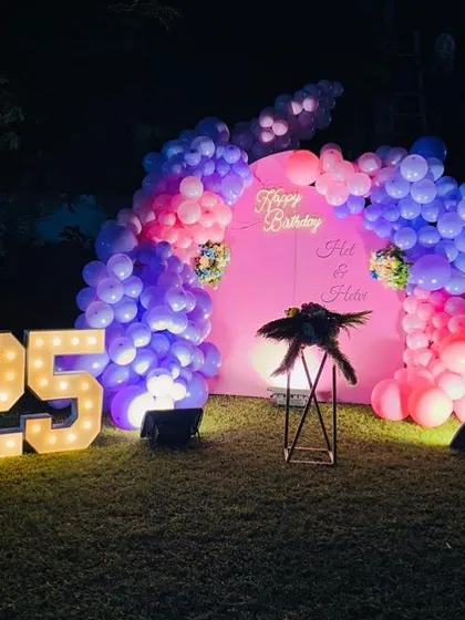A beautiful outdoor setup for a 25th birthday. The decoration features a pink backdrop with a balloon arch in shades of pink and purple, complemented by large marquee numbers.