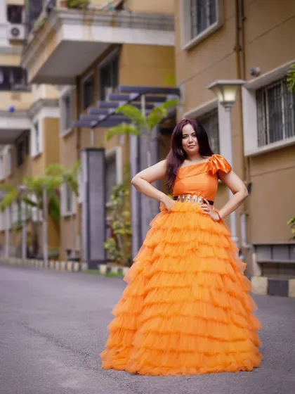A full-length shot of the orange ruffle gown. The one-shoulder design and belted waist create a flattering and stylish silhouette.