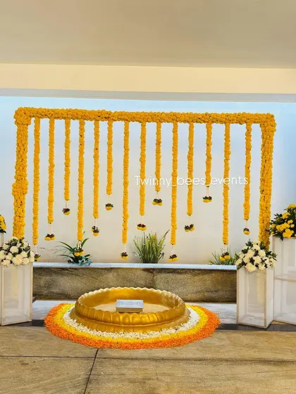 A clean and modern Haldi setup with a marigold arch against a white wall. The large brass urli is placed on a rangoli made of flower petals, combining traditional and contemporary elements.