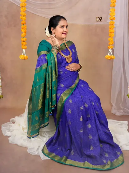A graceful solo portrait. The mom-to-be looks elegant in her blue and green silk saree, seated against a simple backdrop with festive floral garlands.