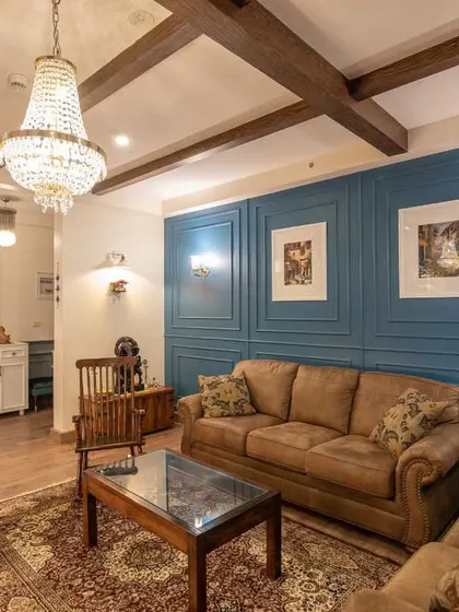 A classic drawing room setup featuring a rich brown chesterfield sofa set against a dusty blue accent wall with decorative molding. The crystal chandelier and traditional rug add a layer of Victorian elegance.