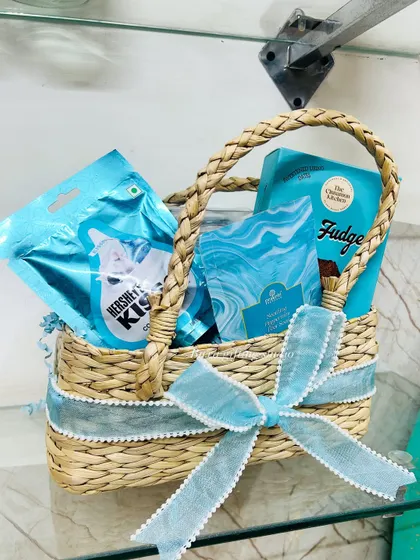 A close-up of the new mom hamper, showing the handwoven basket filled with blue-themed goodies like Hershey's Kisses and The Cinnamon Kitchen fudge.