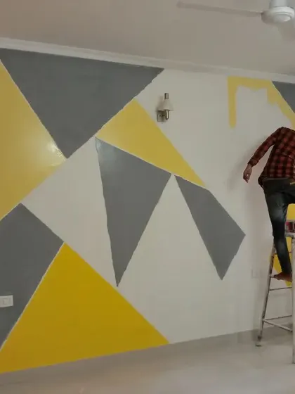 A painter at work on a geometric accent wall in a bedroom, carefully applying shades of yellow, grey, and white to create a bright and modern pattern.