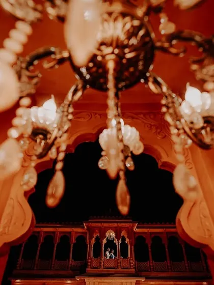 A dramatic, wide-angle pre-wedding shot. The couple stands in a grand, orange-lit archway at night, with a chandelier adding to the opulence.