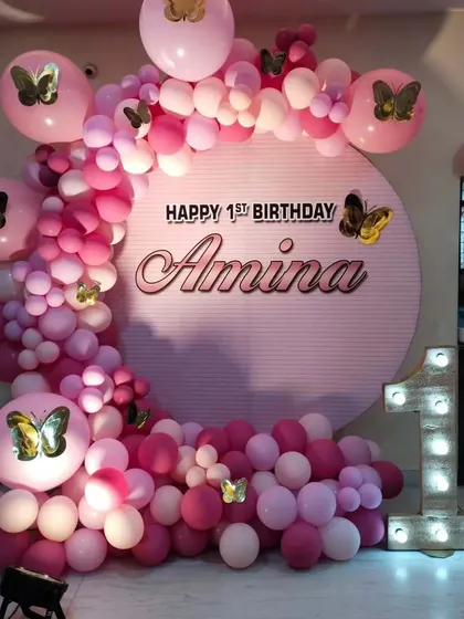 A pink butterfly-themed first birthday. The circular backdrop with the child's name is surrounded by a pink balloon arrangement, butterfly stickers, and a large marquee number '1'.