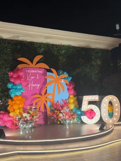 A vibrant tropical theme for a 50th birthday bash. The setup includes a green wall, colorful backdrops with palm tree cutouts, and a large marquee number.
