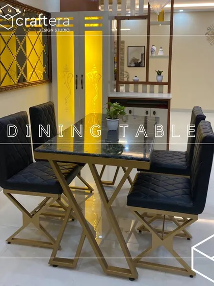 A modern dining setup featuring a black marble-top table with a striking gold-finished base and quilted black leatherette chairs. The lit pooja unit and mirrored wall in the background add to the room's luxurious feel.