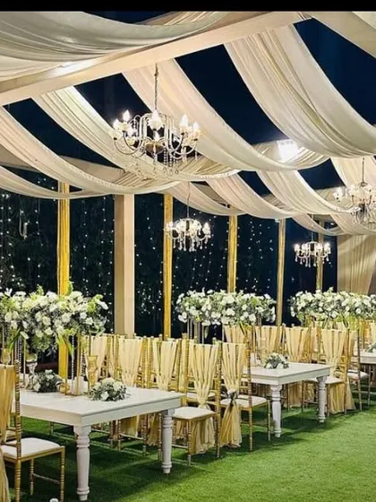 This elegant outdoor dining setup is perfect for a sophisticated reception. It features white tables and chairs with gold sashes, set on an artificial grass carpet under a ceiling of white drapes and chandeliers.