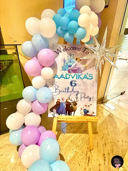 A personalized welcome sign for Aadivka's 6th birthday party. The sign, featuring characters from Frozen, is placed on an easel and framed with a small balloon garland to match the party's theme.