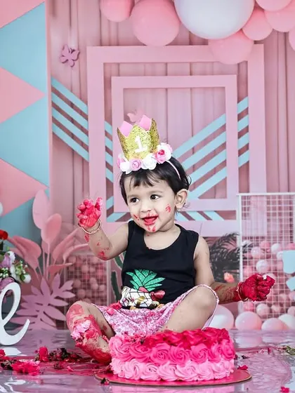 A little boy enjoys his pink-themed cakesmash, with frosting on his hands and a happy expression. The elaborate pink and blue geometric background shows the level of detail in my themed setups.
