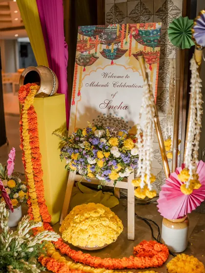 A welcome corner for a Mehendi ceremony, featuring a custom sign, colorful paper fans, and a playful overflow of marigold flowers from a traditional pot.