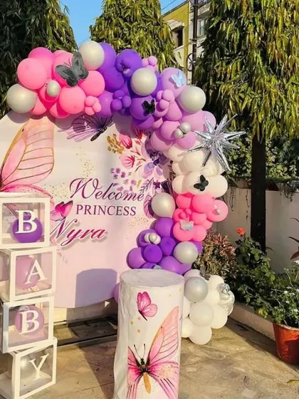 An elegant outdoor setup to "Welcome Princess Nyra". The butterfly-themed decoration includes a personalized round backdrop, a purple and pink balloon arch, and "BABY" blocks.