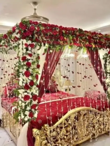 I create luxurious and romantic first night room decorations. This opulent setup features a four-poster bed draped in red fabric and completely covered in fresh red and white roses, with delicate floral strings hanging down for a magical feel.