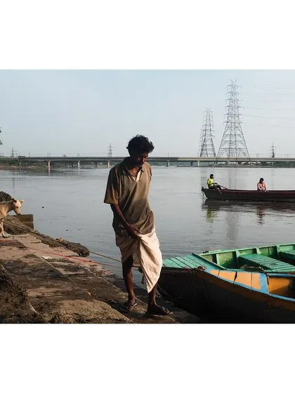 A quiet moment on the banks of the Yamuna, with a boatman standing by his boats and a dog walking along the ghat. In the background, another boat drifts by, completing the tranquil scene.