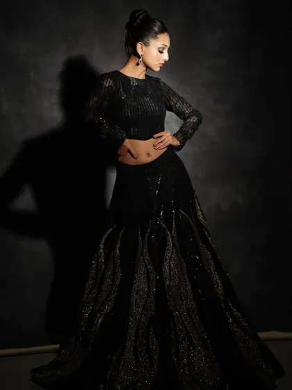 This black fusion lehenga is all about drama and texture. The skirt features unique, wave-like sculpted details, paired with a sleek, long-sleeved sequin crop top for a look that is both edgy and sophisticated.