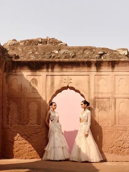 Two models styled in ivory outfits for a campaign shoot. The setting, with its weathered walls and clean pink backdrop, creates a modern yet timeless feel that complements the elegant designs.