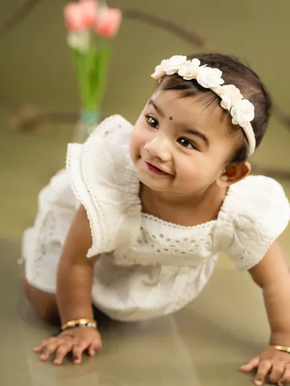 A baby girl in a white dress and floral headband, crawling towards the camera with a sweet smile. A classic and heartwarming baby portrait.