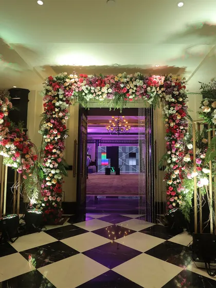 A grand floral entrance leading into a banquet hall with a striking black and white checkered floor. The abundance of pink, red, and white flowers creates a stunning contrast and a luxurious welcome.