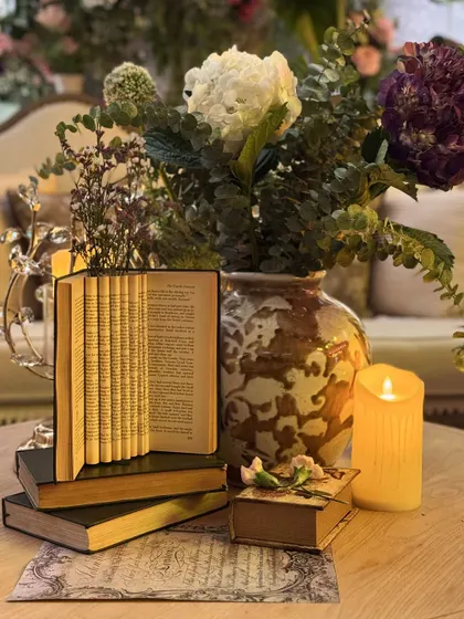 A table centerpiece that is a story in itself. A stack of vintage books, one artfully folded, is paired with a single candle and a floral arrangement, creating a poetic and literary-inspired detail.