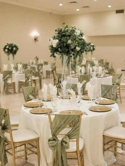 An indoor reception hall with round tables and Chiavari chairs decorated with sage green sashes. The tall floral centerpieces add height and drama to the room.
