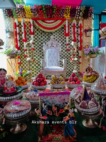 A grand Seemantham setup featuring a silver throne and a checkerboard backdrop in red, yellow, and white. The stage is filled with an elaborate display of traditional offerings and dolls.